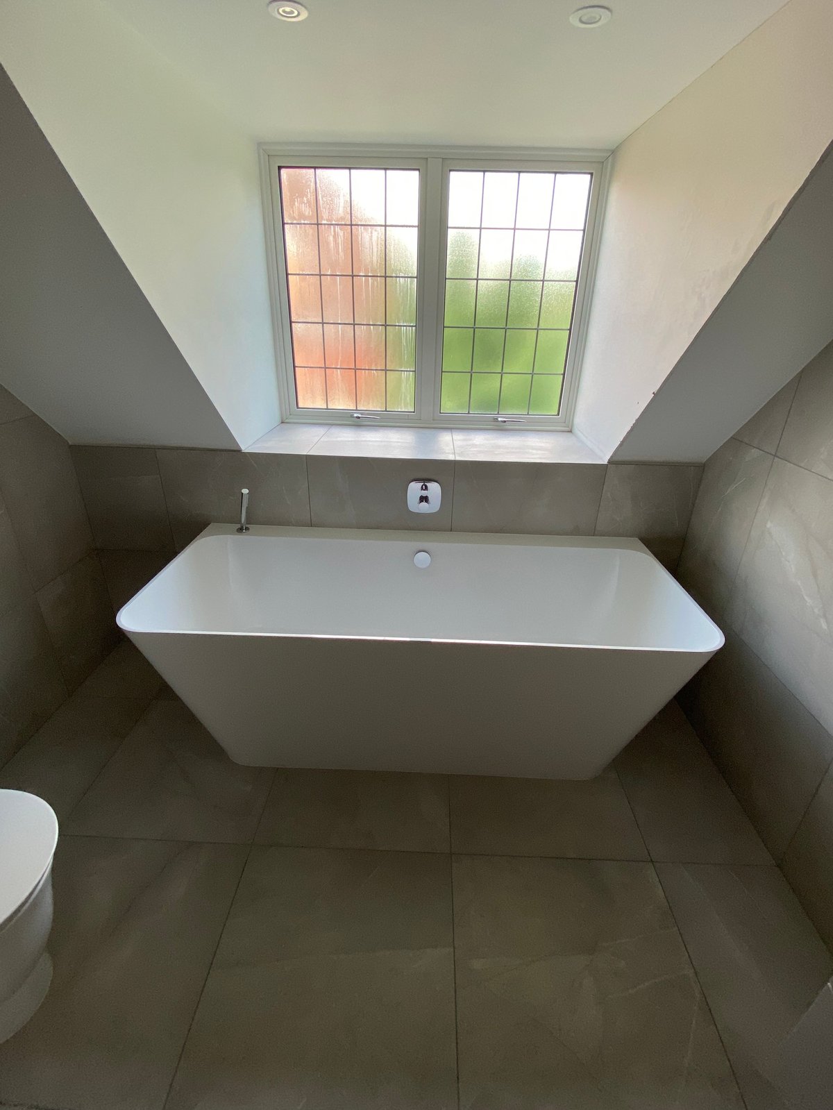 Modern bathroom with white freestanding bathtub, gray tile walls and floors, and skylight window with colorful stained glass above.