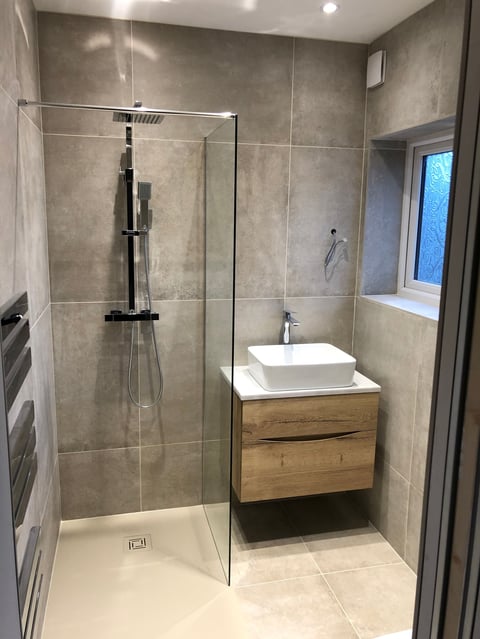 Modern bathroom with walk-in shower enclosure, white vessel sink on wooden vanity, gray tile walls, and recessed lighting