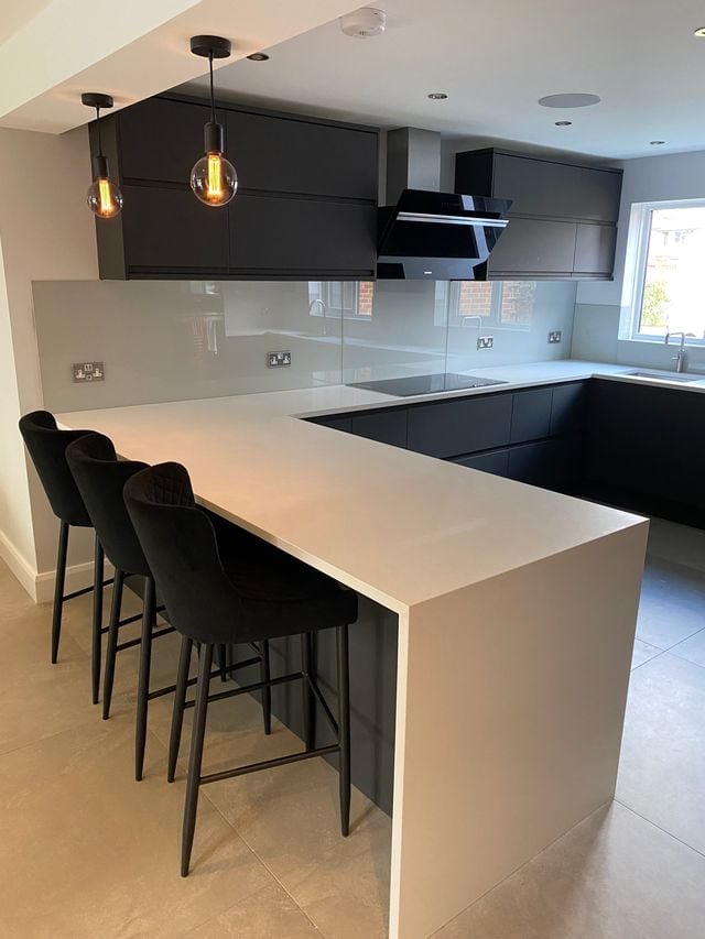 Modern kitchen with black upper cabinets, white countertop island, black bar stools, and glass backsplash with pendant lighting
