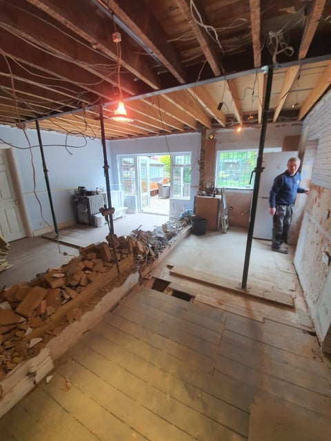 Interior of a residential space under renovation with exposed ceiling beams, debris on wooden floor, and a worker in the background near glass doors.