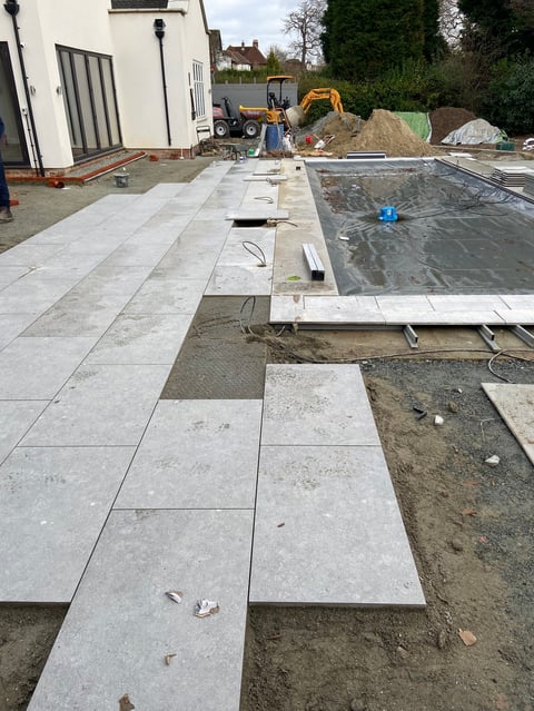 Backyard patio under construction with gray stone tiles being laid, excavator and construction materials visible in background