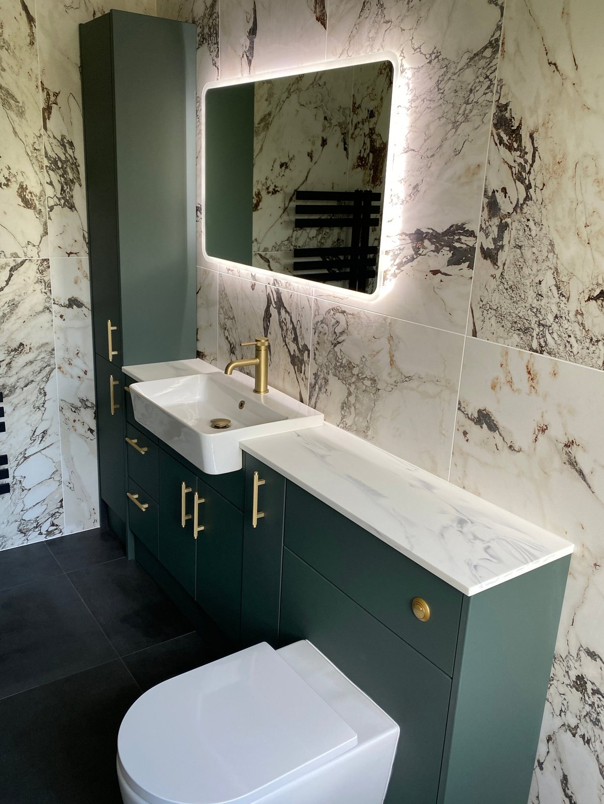 Modern bathroom with green vanity, white sink, illuminated mirror, marble-pattern walls, and white toilet on dark floor.
