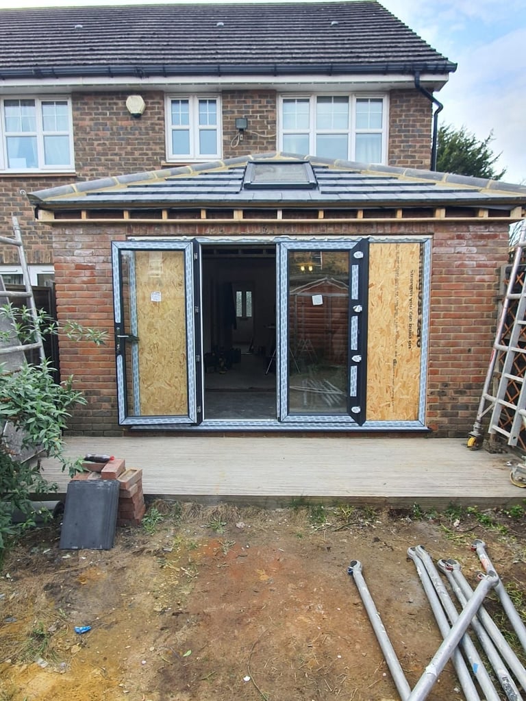 Brick house with modern glass extension under construction, featuring boarded windows and new patio foundation