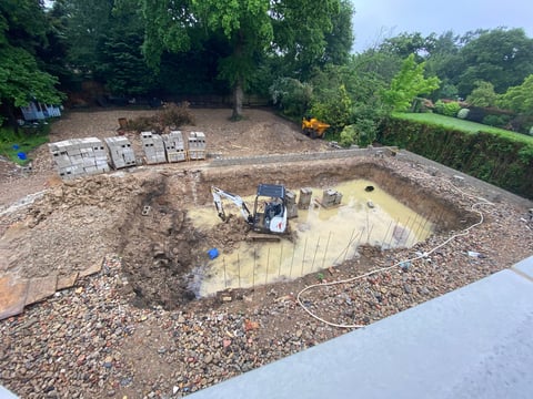 Construction site with excavator in muddy pit surrounded by gravel, building materials, and green trees