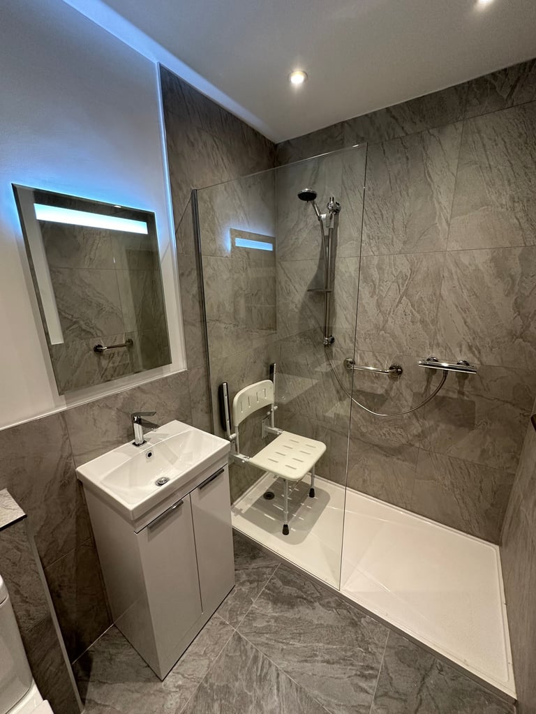 Modern bathroom with gray marble tiles, white vanity sink, illuminated mirror, and walk-in shower with glass enclosure