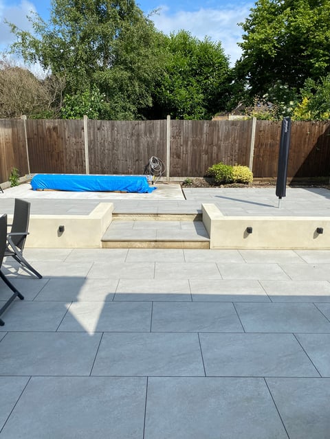 Patio with blue pool cover, sunken seating area, potted plant, wooden fence, and trees in background on sunny day