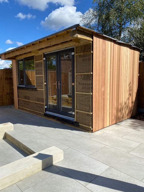 Modern wooden garden shed with glass doors and windows on a paved patio under clear sky
