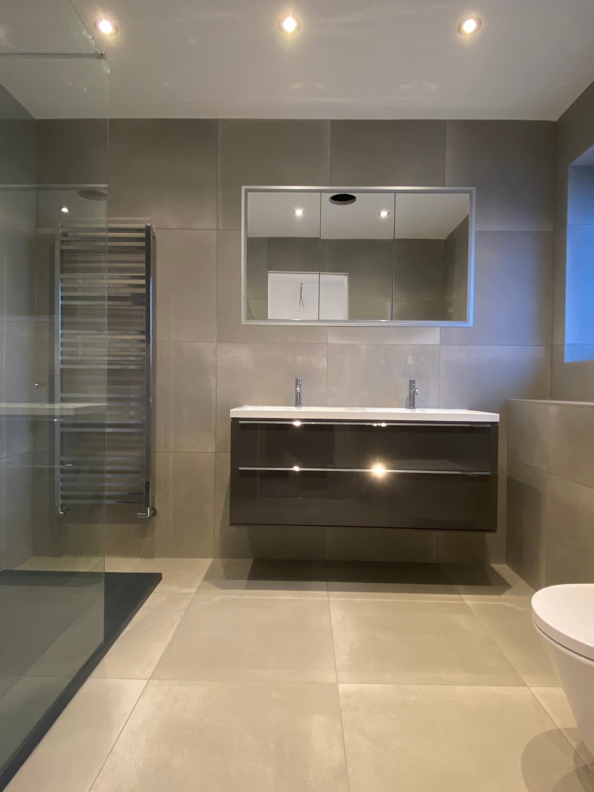 Modern bathroom with dark vanity cabinet, dual sinks, large mirror, glass shower enclosure, and neutral tile flooring