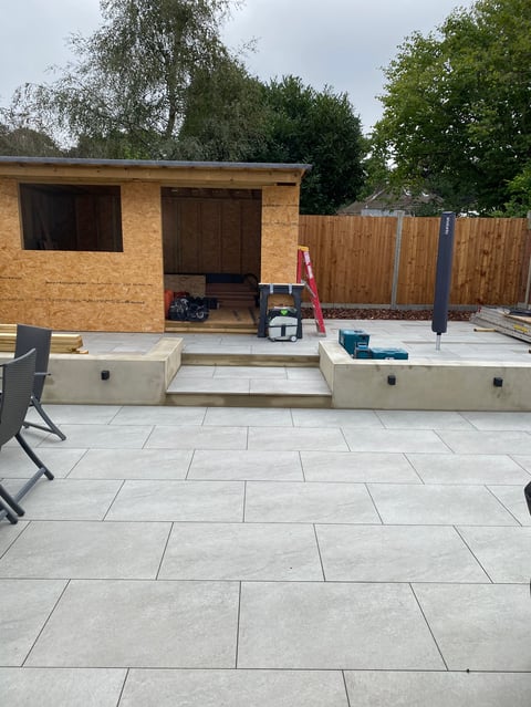 Construction site with a brick garden shed, raised platform, paved patio, and wooden fence under renovation