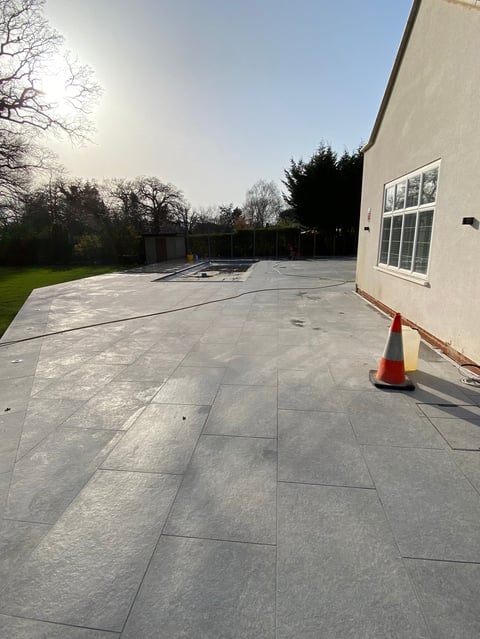 New gray patio with large tiles in a residential backyard under construction with a red traffic cone and modern house corner visible