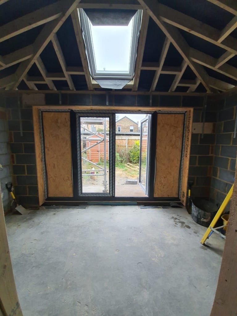 Interior of a room under construction with exposed wooden beam ceiling, skylight, and black-framed bifold doors overlooking a garden