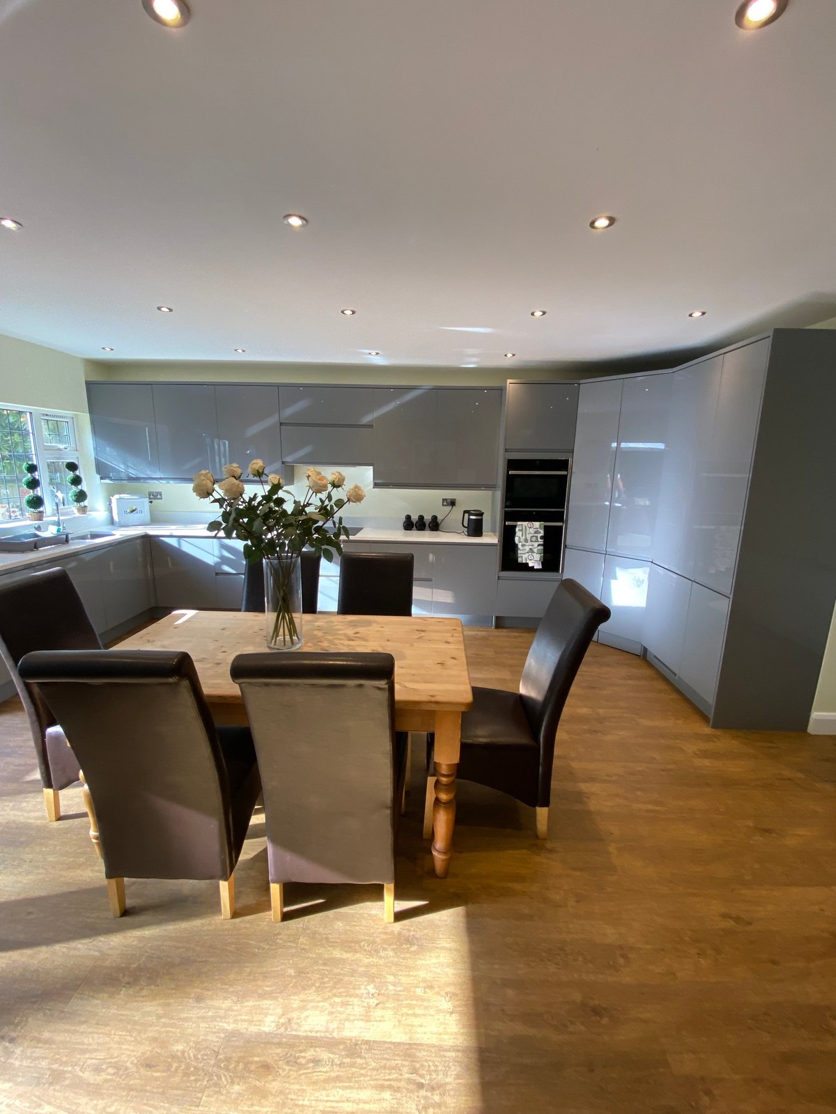 Modern open-plan kitchen with wooden dining table, gray upholstered chairs, white cabinetry, and recessed lighting