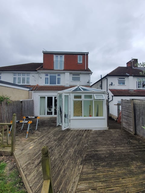 White conservatory with glass panels on a wooden deck extending from a brick home, surrounded by neighboring houses under cloudy sky