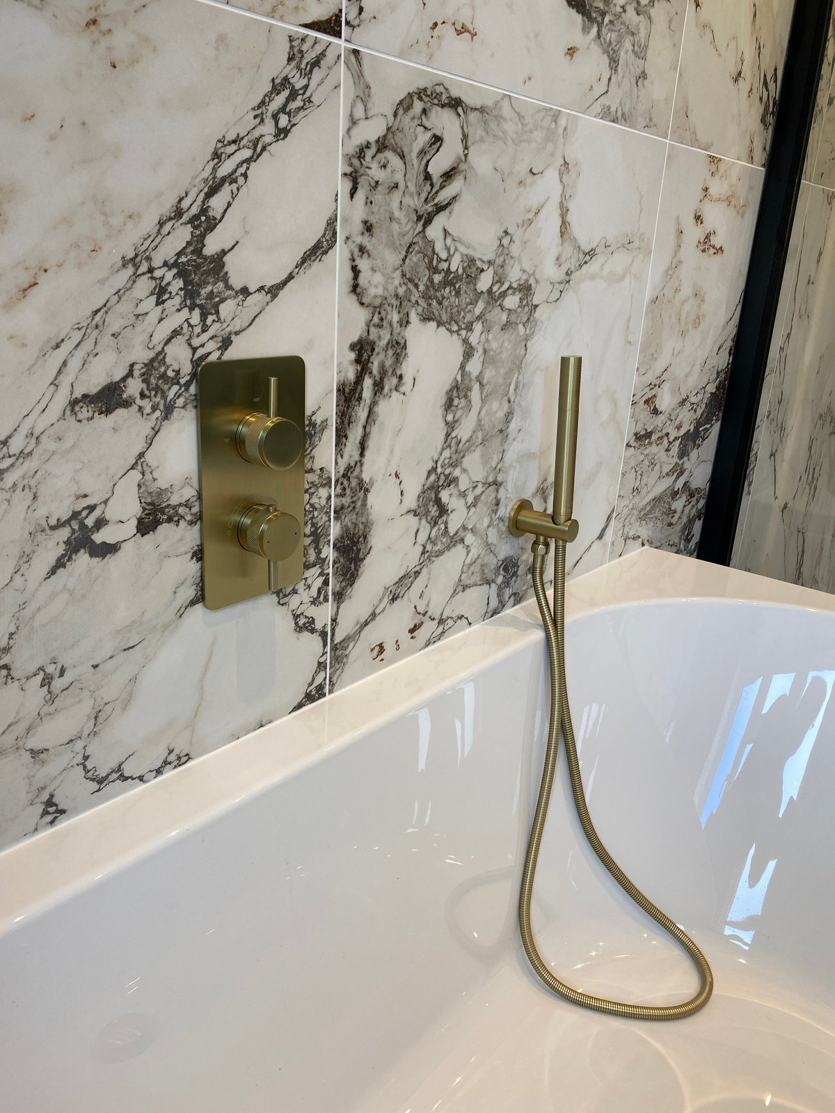 Modern bathroom tub with marble tile walls and brass shower fixtures with handheld spray
