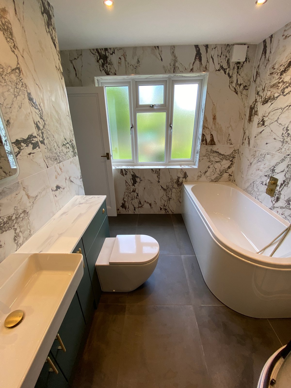 Modern bathroom with marble-patterned walls, white fixtures including a toilet and curved bathtub, sink with green vanity, and window overlooking garden