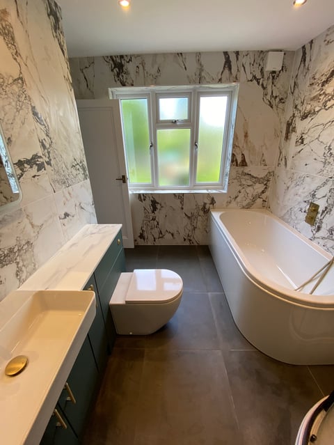 Modern bathroom with marble-patterned walls, white fixtures including a toilet and curved bathtub, sink with green vanity, and window overlooking garden