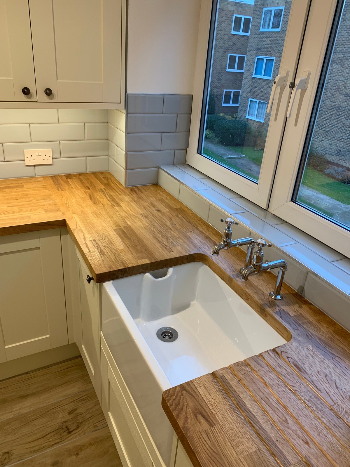 Modern kitchen with white cabinets, wooden countertop, white undermount sink with chrome faucet, and large window overlooking residential building.