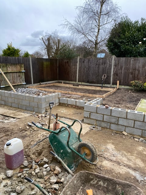 A green wheelbarrow in a residential backyard garden under construction with raised wooden beds and wooden fencing