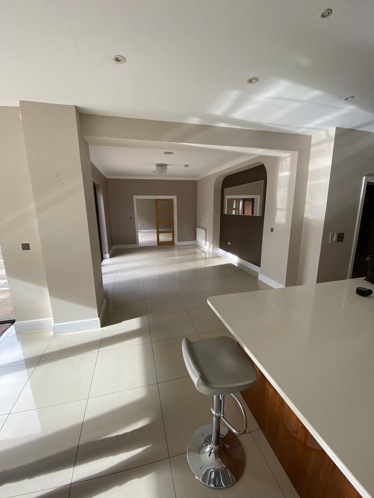 Bright modern open-plan home interior with white and beige walls, tiled flooring, bar stool at counter, and multiple doorways with natural light