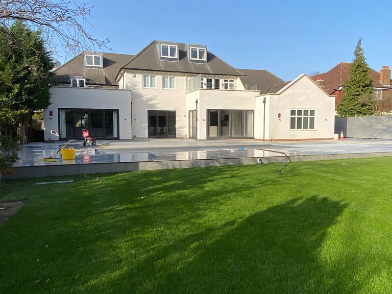 Modern two-story house with cream exterior, dark roof, large lawn, swimming pool, and landscaped garden on sunny day